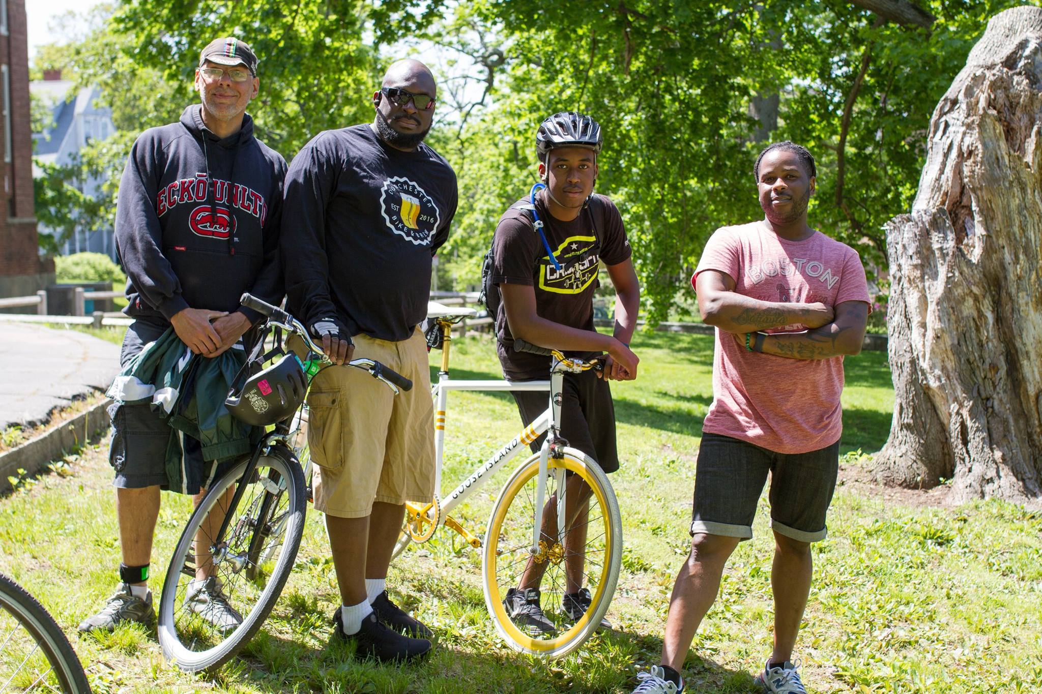An image of 4 men of different ages standing around 2 bikes, in a park with greenery around them.